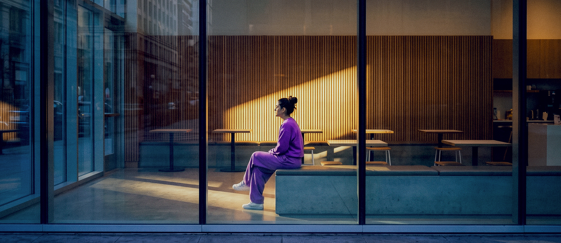 Persona con suéter morado sentada en un espacio moderno con ventanas de cristal, vista a la ciudad y luz natural dorada.