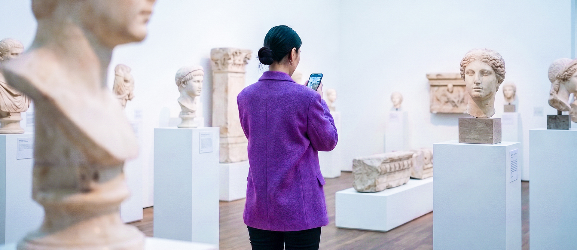 Persona con chamarra morada usando su celular en un museo rodeada de estatuas antiguas, ilustrando la historia de la banca digital: evolución de ventanillas y filas a apps 100% digitales en tu celular.