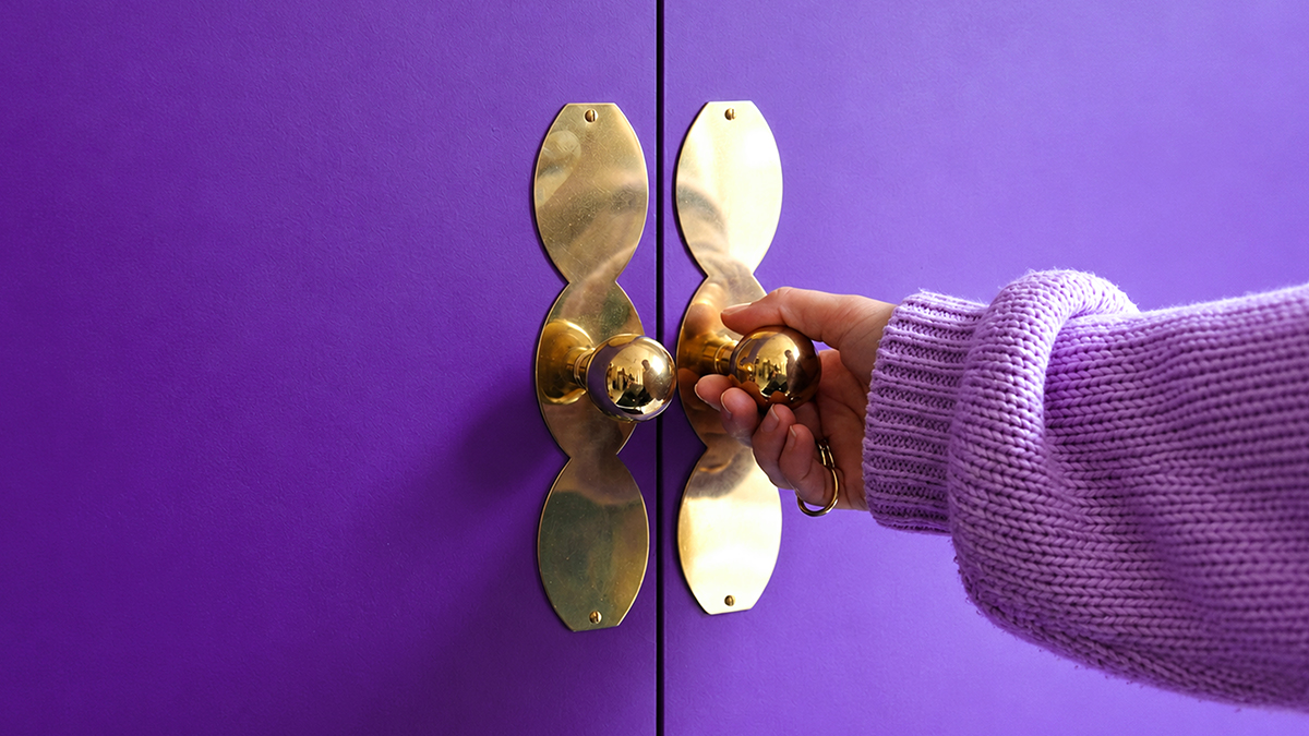 Mano con suéter morado abre unas puertas moradas con manijas doradas, una metáfora de cómo organizar tus finanzas en la app Nu con unos pocos ajustes.