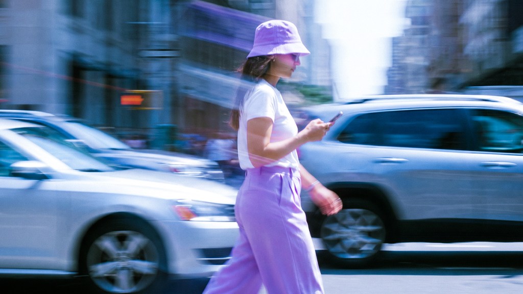 Mujer con sombrero y pantalón morado camina con celular en mano mientras los autos pasan.