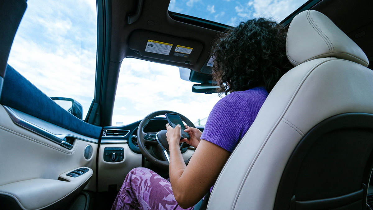 Mujer sentada al volante de un auto consulta qué puede hacer desde la app Nu en su celular, con el cielo de fondo.