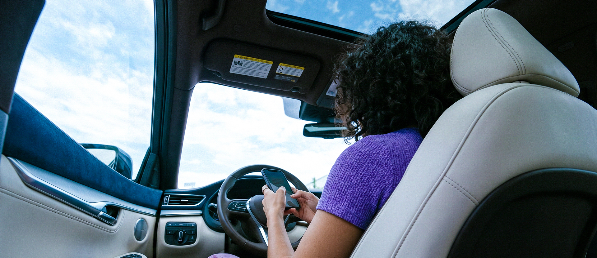 Mujer sentada al volante de un auto consulta qué puede hacer desde la app Nu en su celular, con el cielo de fondo.