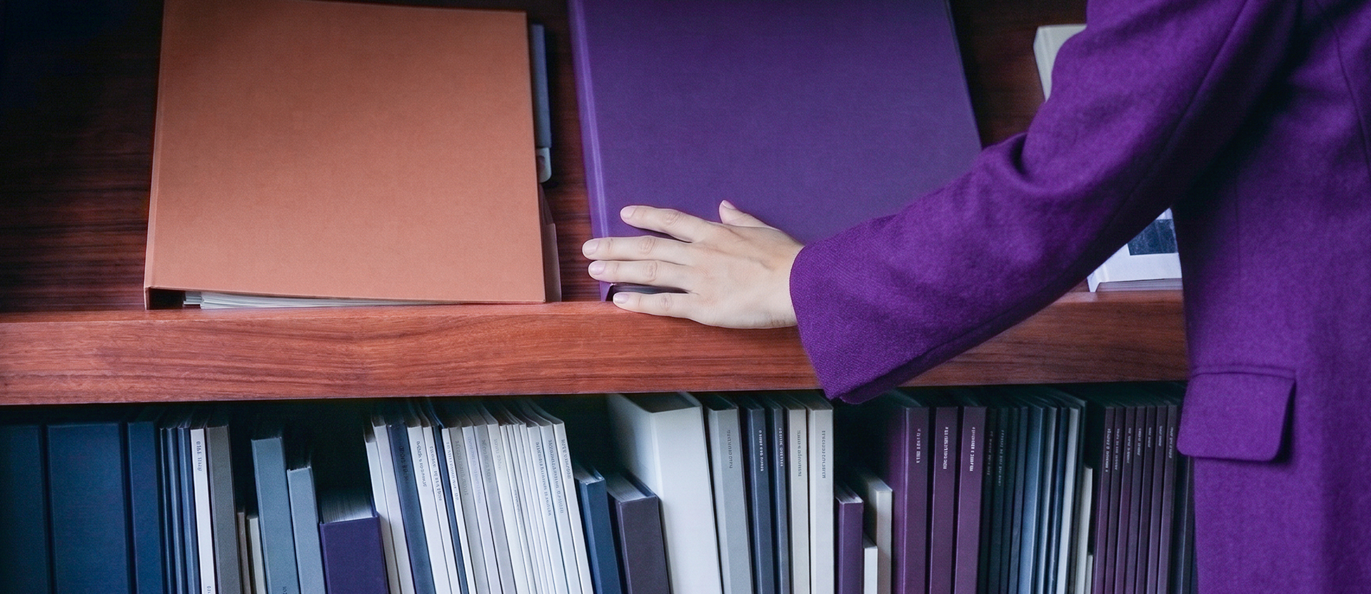 Mano de una persona con abrigo morado sobre una carpeta morada en un librero de madera lleno de archivos.