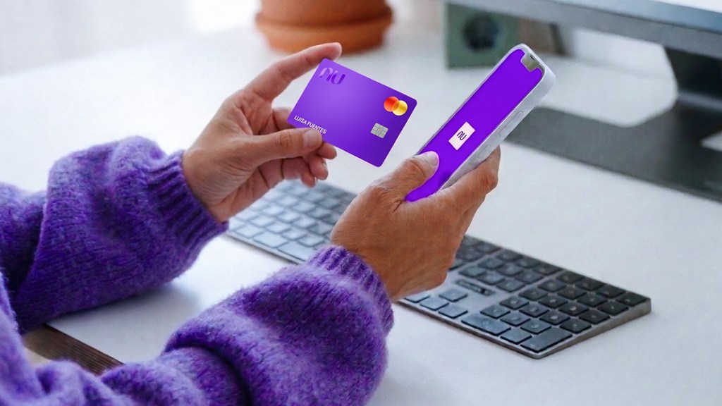 Manos con suéter morado sostienen una tarjeta Nu y un celular con la app abierta, para elegir entre tarjeta de débito virtual vs física según cada compra.