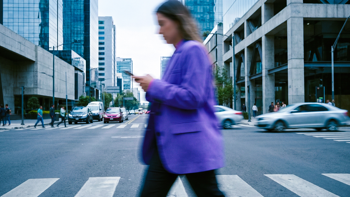 Mujer con blazer morado cruza una calle mientras revisa su celular, una escena cotidiana donde conocer los tipos de phishing ayuda a evitar fraudes.